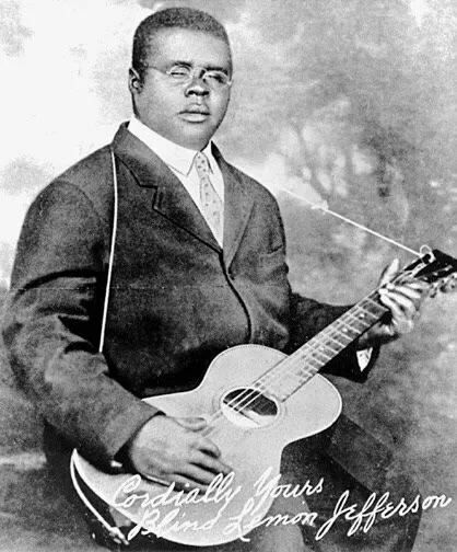 Black-and-white publicity portrait of Blind Lemon Jefferson seated with a guitar and wearing a dark suit, round glasses, and tie.