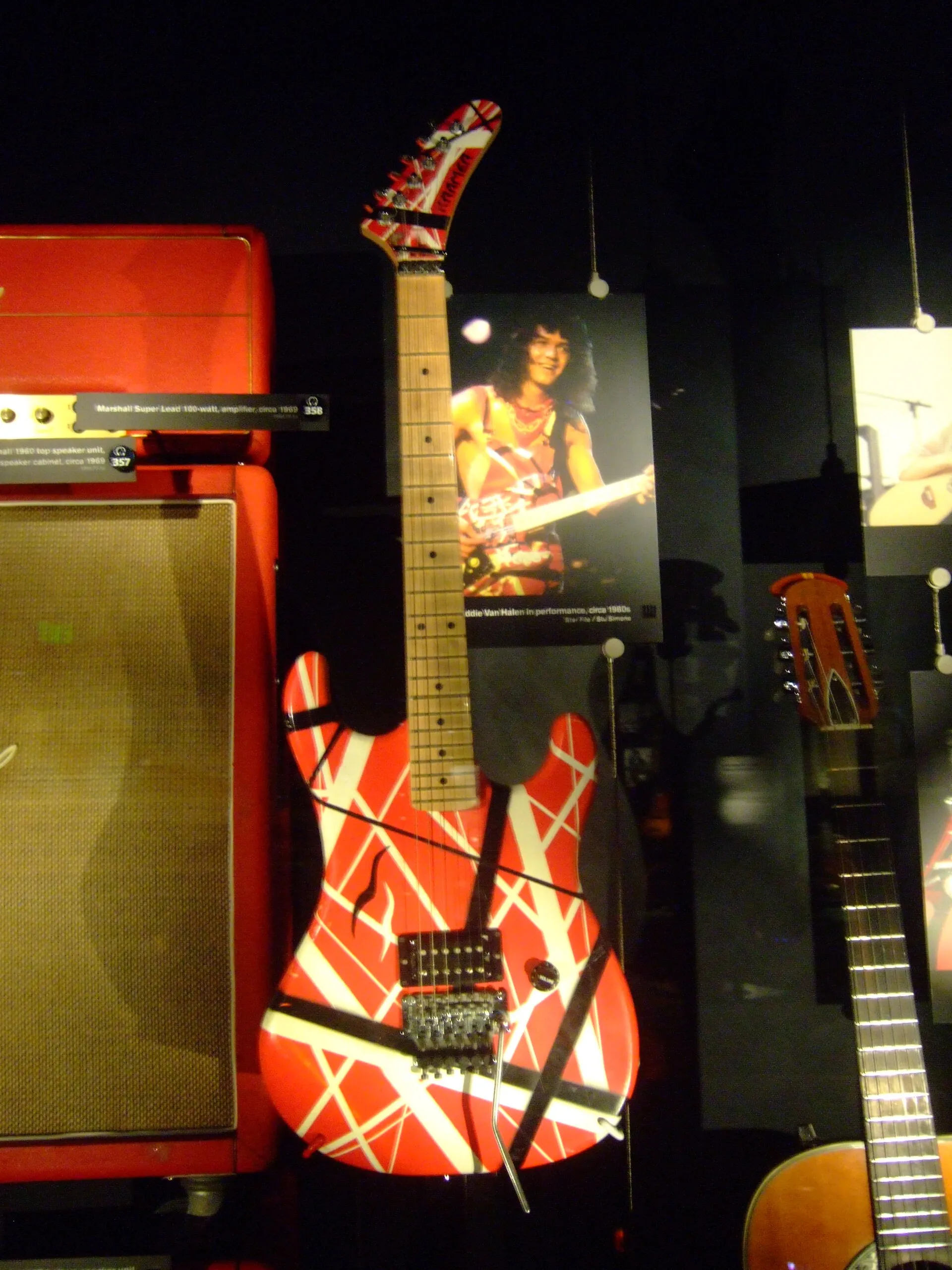 Red, white, and black striped Eddie Van Halen electric guitar displayed upright beside Marshall amplifier cabinets in a museum.