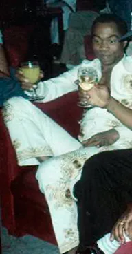 Fela Kuti stands in profile in 1970, wearing patterned trousers and holding a glass beside another man.