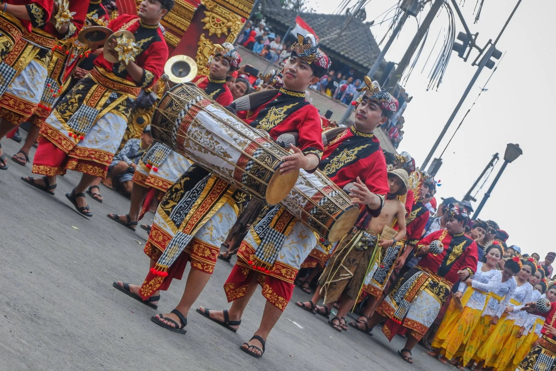A drummer in red ceremonial dress plays a long double-headed kendang while walking at the front of a Balinese procession.
