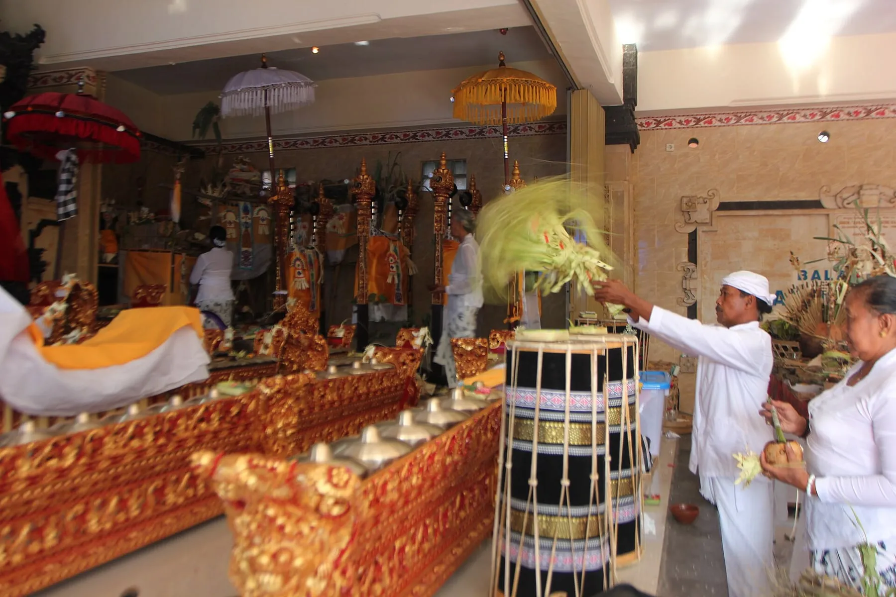 Two men in white ceremonial clothing stand among carved red-and-gold gamelan instruments during a pemalaspasan ceremony in Bali.