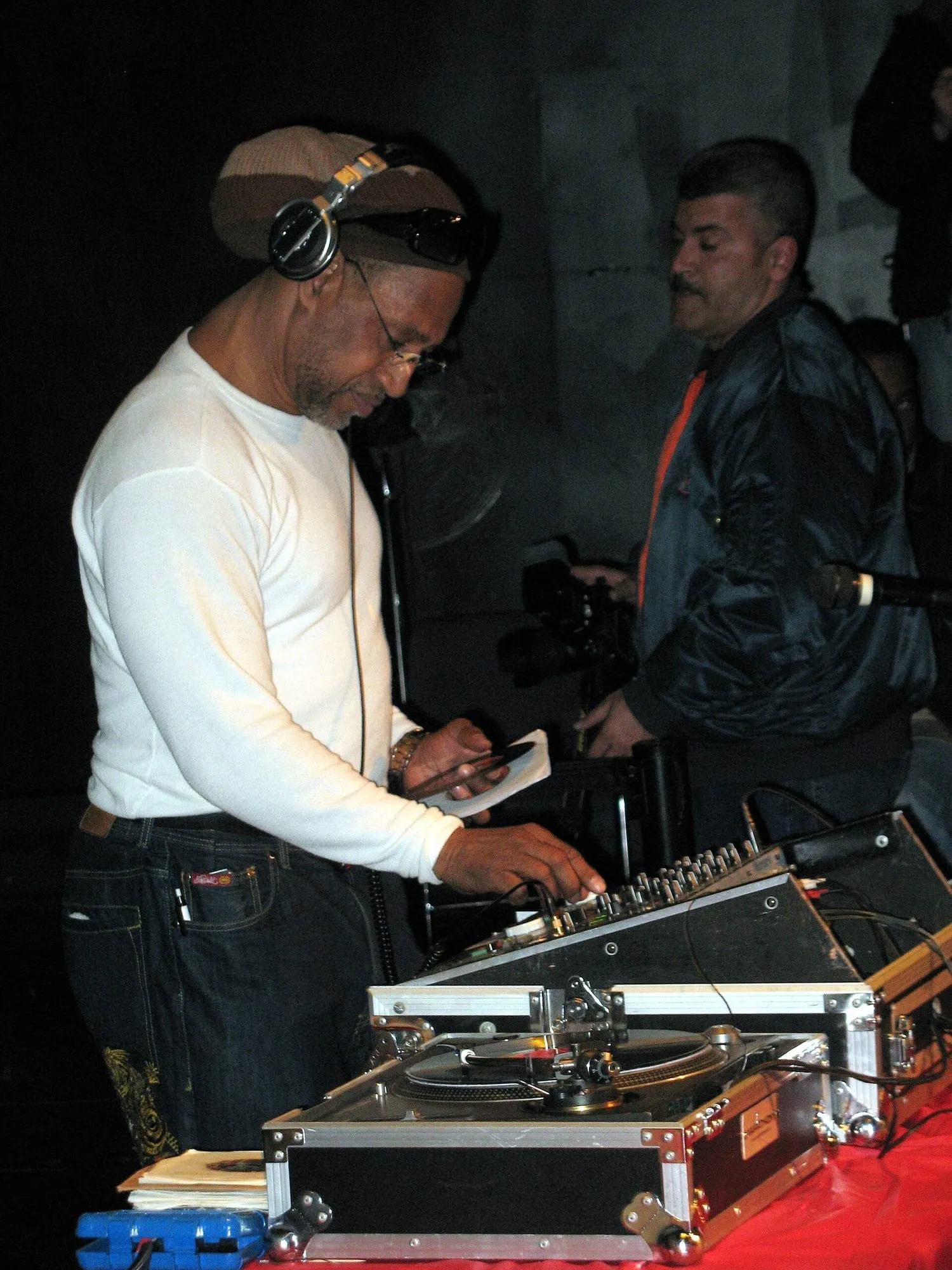 DJ Kool Herc stands behind turntables and a mixer beneath a dark canopy during a 2009 outdoor event in the Bronx.