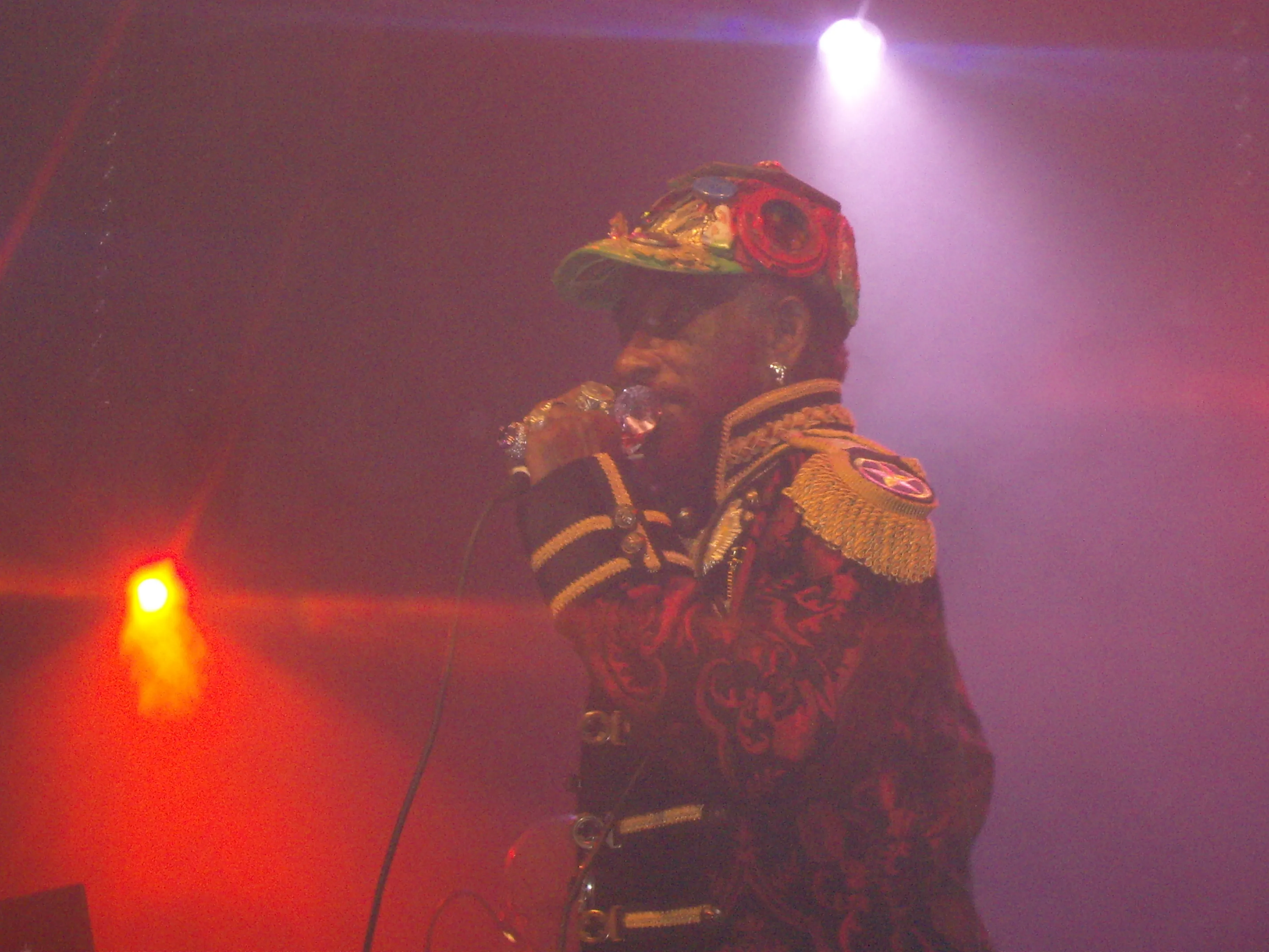 Lee Scratch Perry performs at a festival in Grenoble in 2009, singing into a handheld microphone beneath blue stage light.