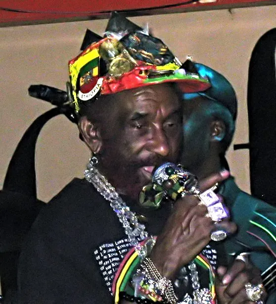 Lee Scratch Perry faces the camera in London in 2008, wearing a decorated cap, mirrored glasses, necklaces, and a patterned jacket.