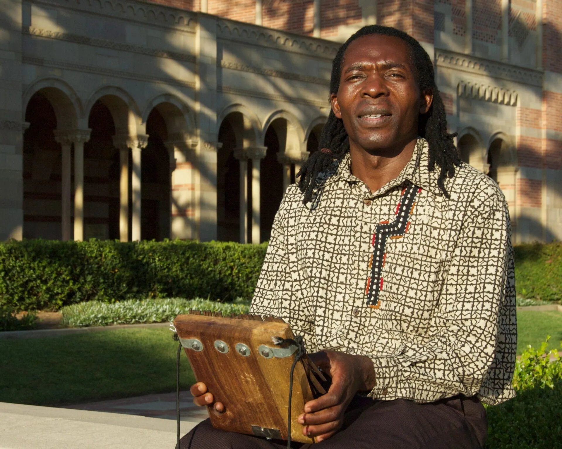 Zimbabwean mbira player Forward Kwenda sits outdoors near UCLA's Royce Hall with the instrument held inside a large gourd resonator.