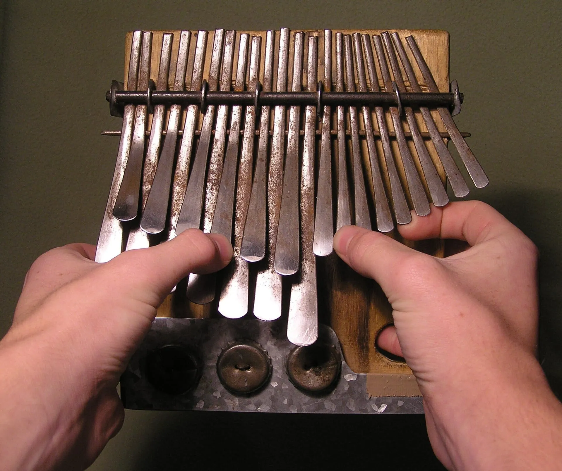 Two hands hold a wooden mbira dza vadzimu and pluck its three banks of closely spaced metal keys with thumbs and one index finger.