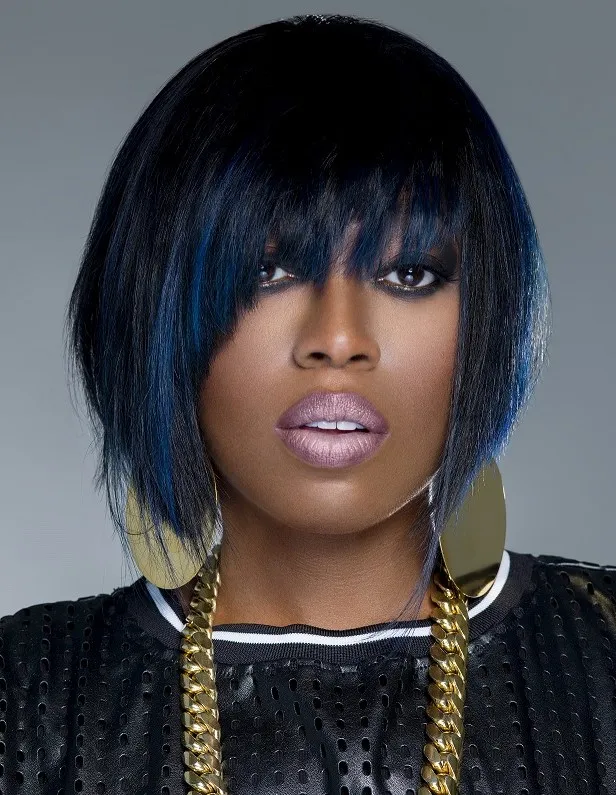 Studio portrait of Missy Elliott facing the camera in a black cap and patterned black-and-white jacket against a pale background.