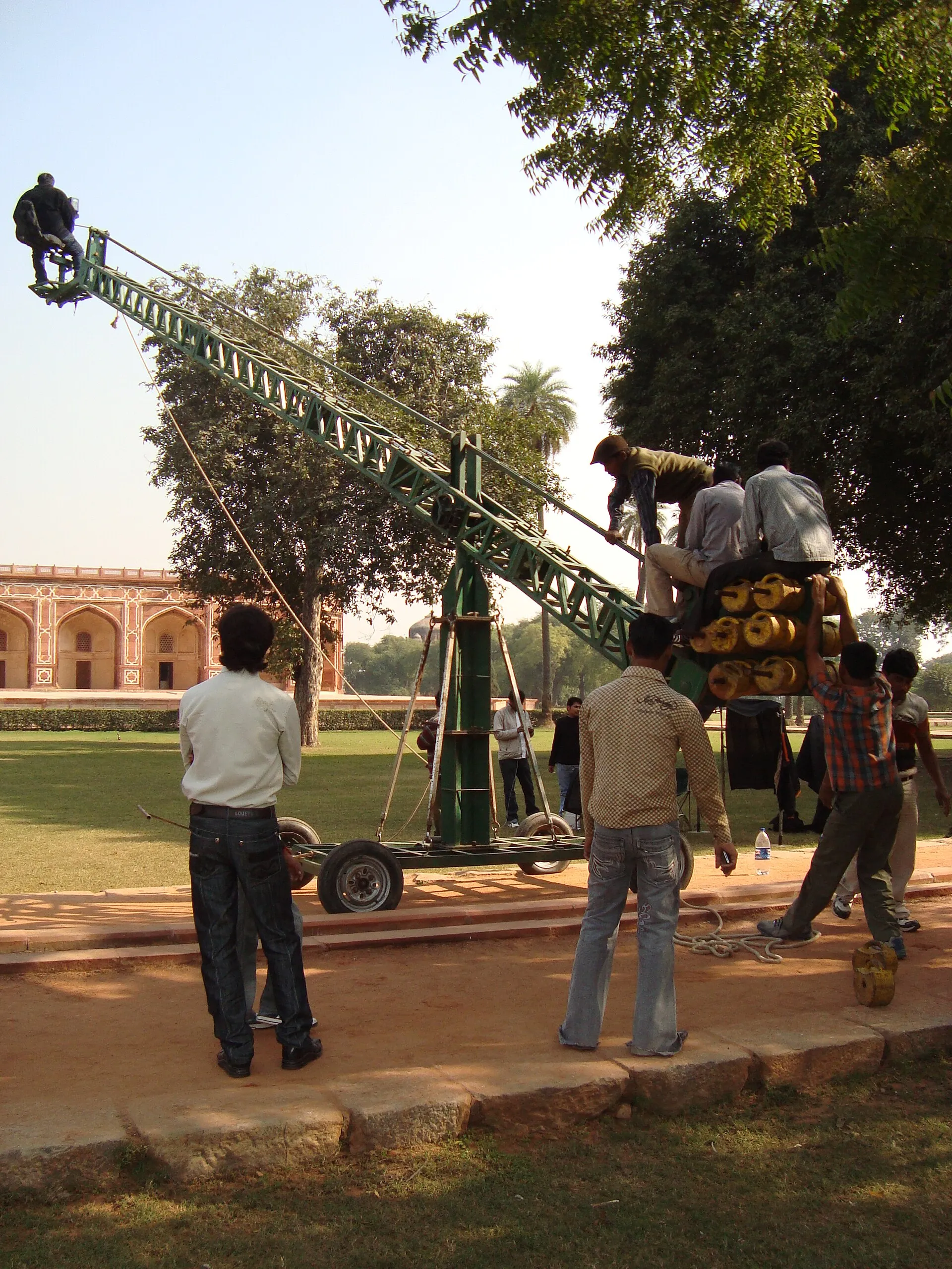 Camera operator seated on a film crane above an outdoor Indian set while crew members work below.
