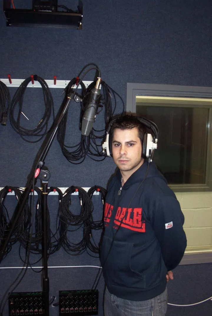 Vocalist wearing headphones and standing beside a large microphone in a separate studio live room.
