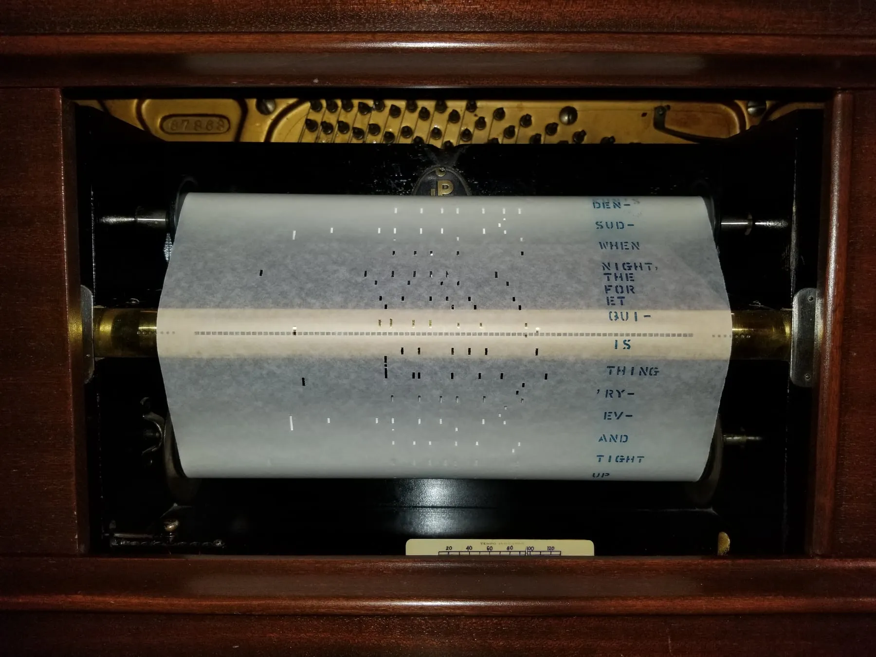 A perforated white piano roll travels across the tracker bar inside a wooden player piano, with printed lyrics and note holes visible on the paper.