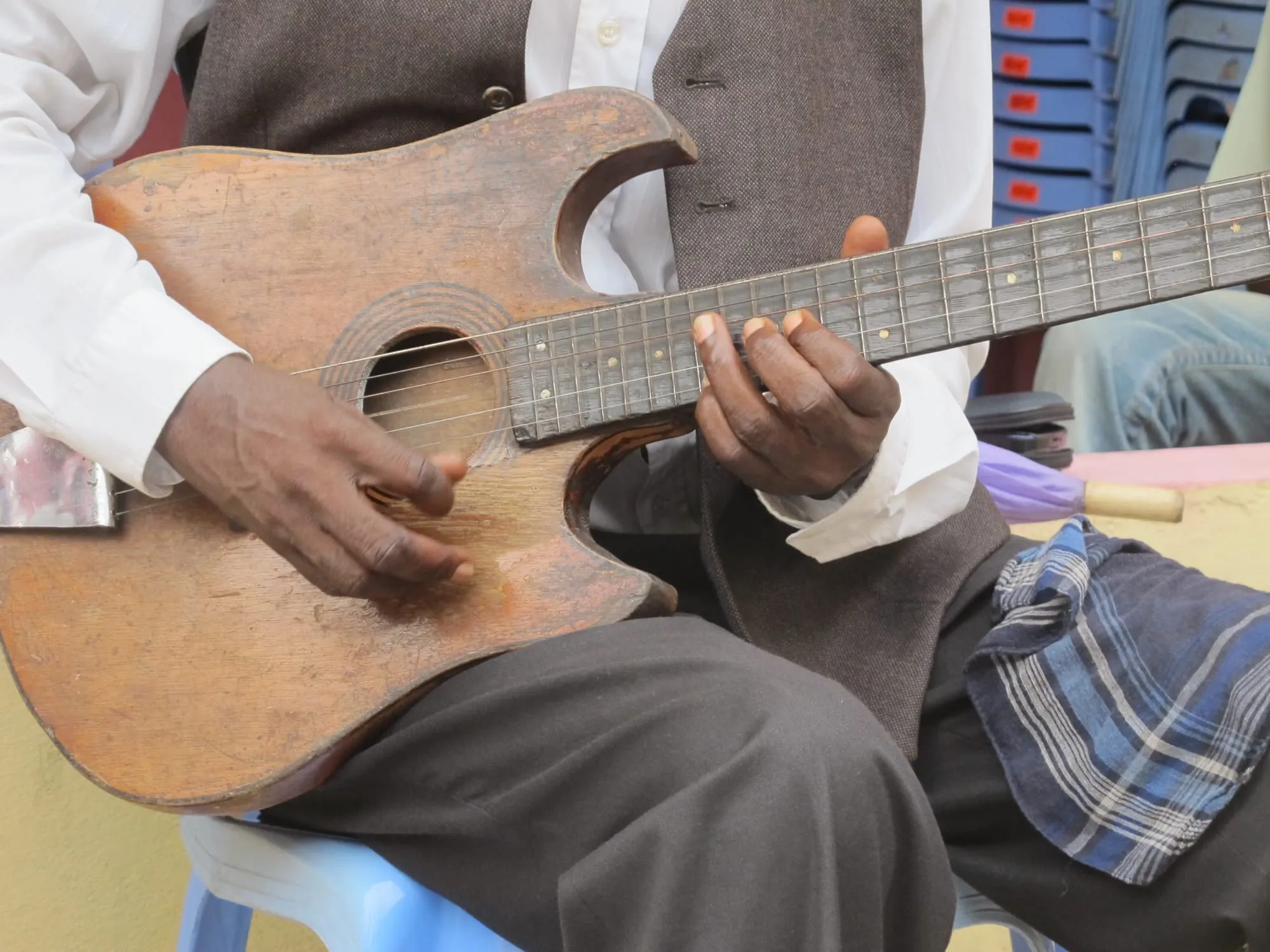 A seated musician's hands play a weathered handmade double-cutaway guitar during a rehearsal in Kinshasa.