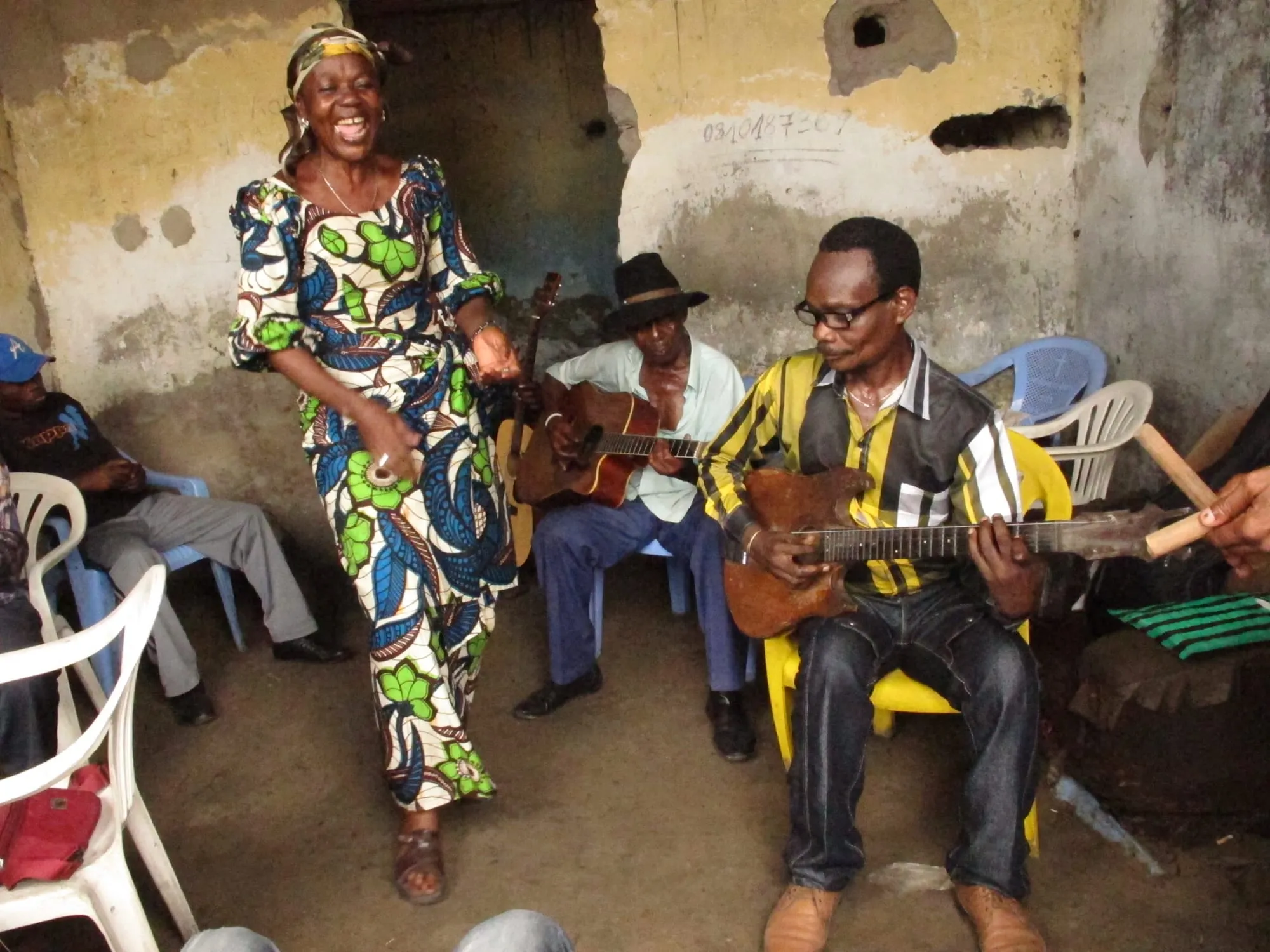 A woman sings beside seated guitarists during a Bakolo Music International rehearsal in a worn Kinshasa room with plastic chairs.