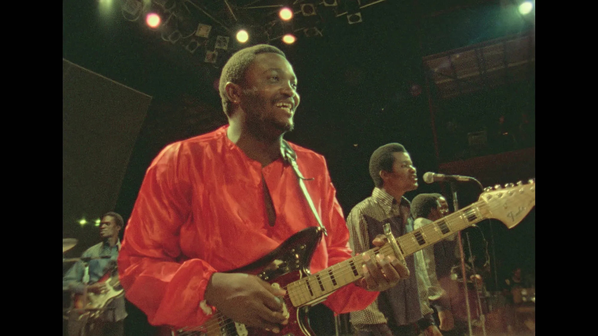 Franco Luambo smiles in a red shirt while playing a black electric guitar with TPOK Jazz onstage at the Zaire 74 festival in Kinshasa.