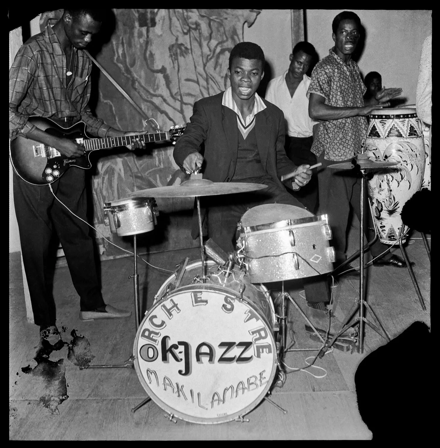 Five members of OK Jazz perform in Leopoldville with electric guitar, compact drums, hand percussion, and a singer gathered in a small room.