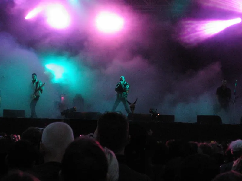 The Sisters of Mercy perform at M'era Luna in 2005 across a wide stage filled with violet, white, and turquoise light and dense haze.