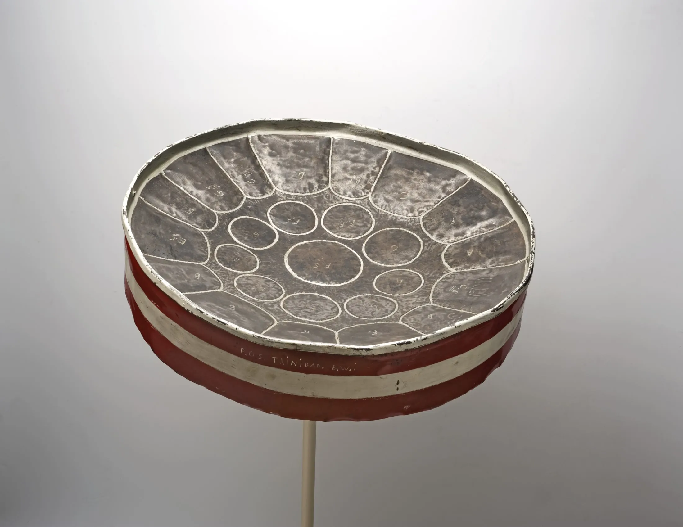 Museum photograph of an early red-and-white tenor steelpan with irregular hand-shaped note areas marked across its worn metal surface.
