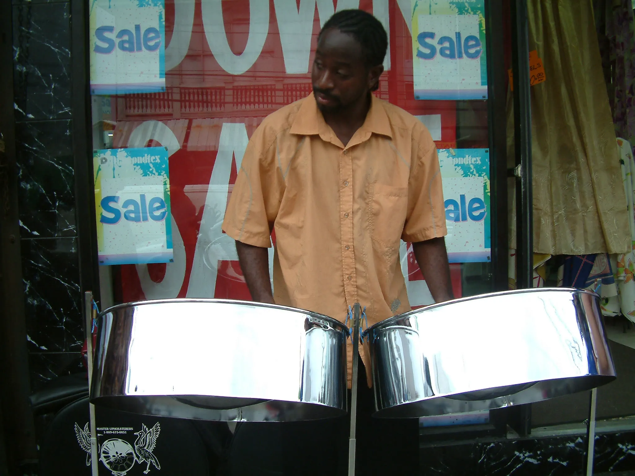 A musician on a street in Trinidad and Tobago looks down while playing two polished steelpans mounted in front of him.