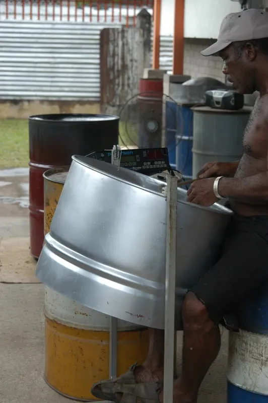A steelpan tuner sits beside several drums and works with hand tools on the playing surface of a silver pan.