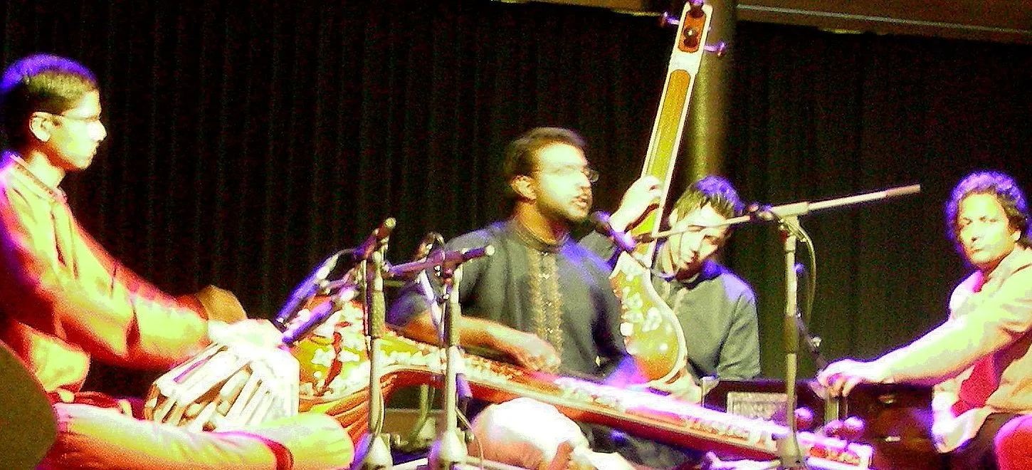 A dhrupad singer performs on a low stage between two tanpura players, with tabla and harmonium players completing the seated ensemble.