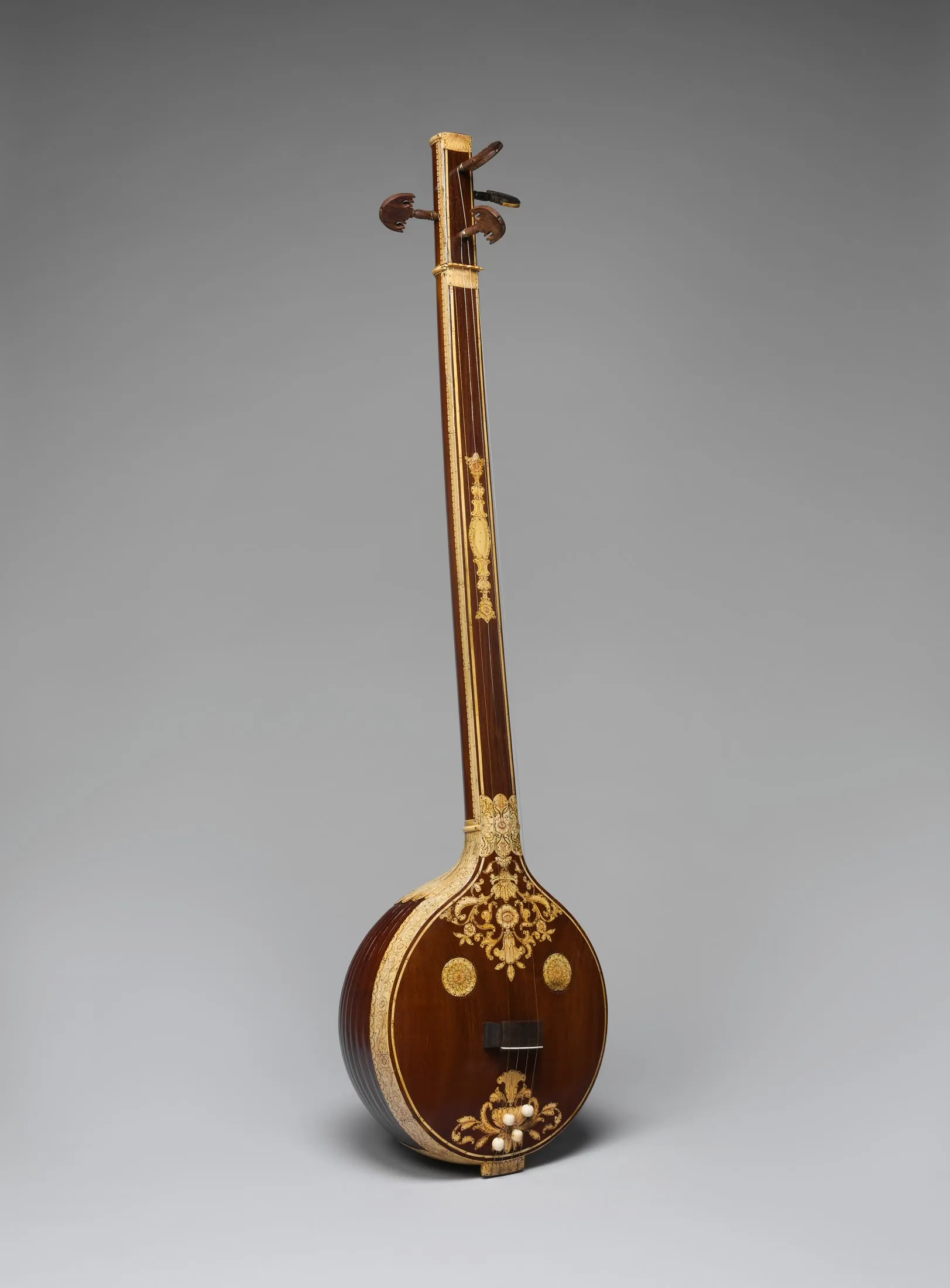 A tall early nineteenth-century Tanjore tanpura with a round wooden resonator, long fretless neck, four pegs, and floral inlay stands against gray.