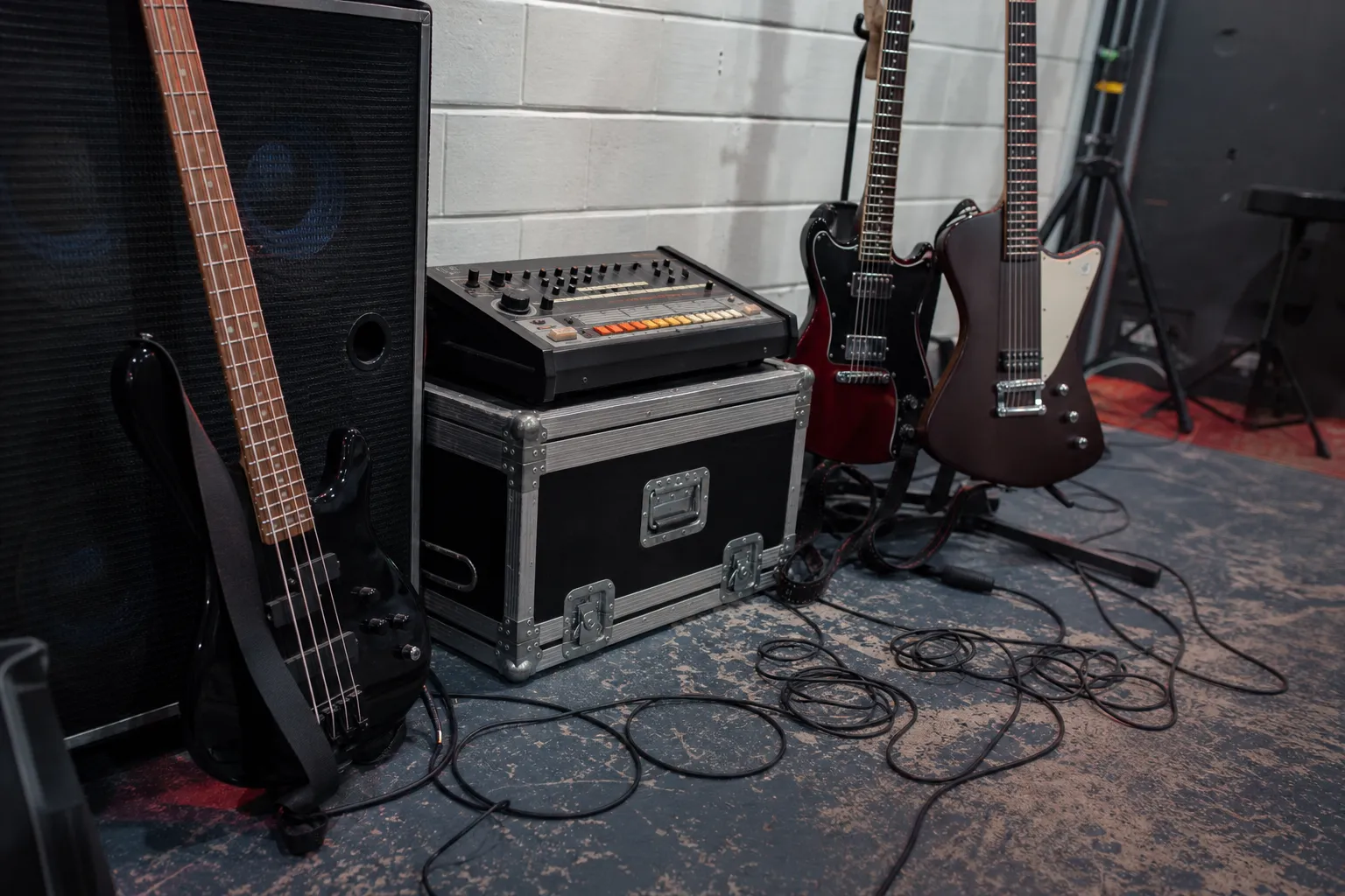 Conceptual editorial photograph of a bass, two electric guitars, a generic early drum machine, amplifier, road case, and cables in an empty rehearsal room.