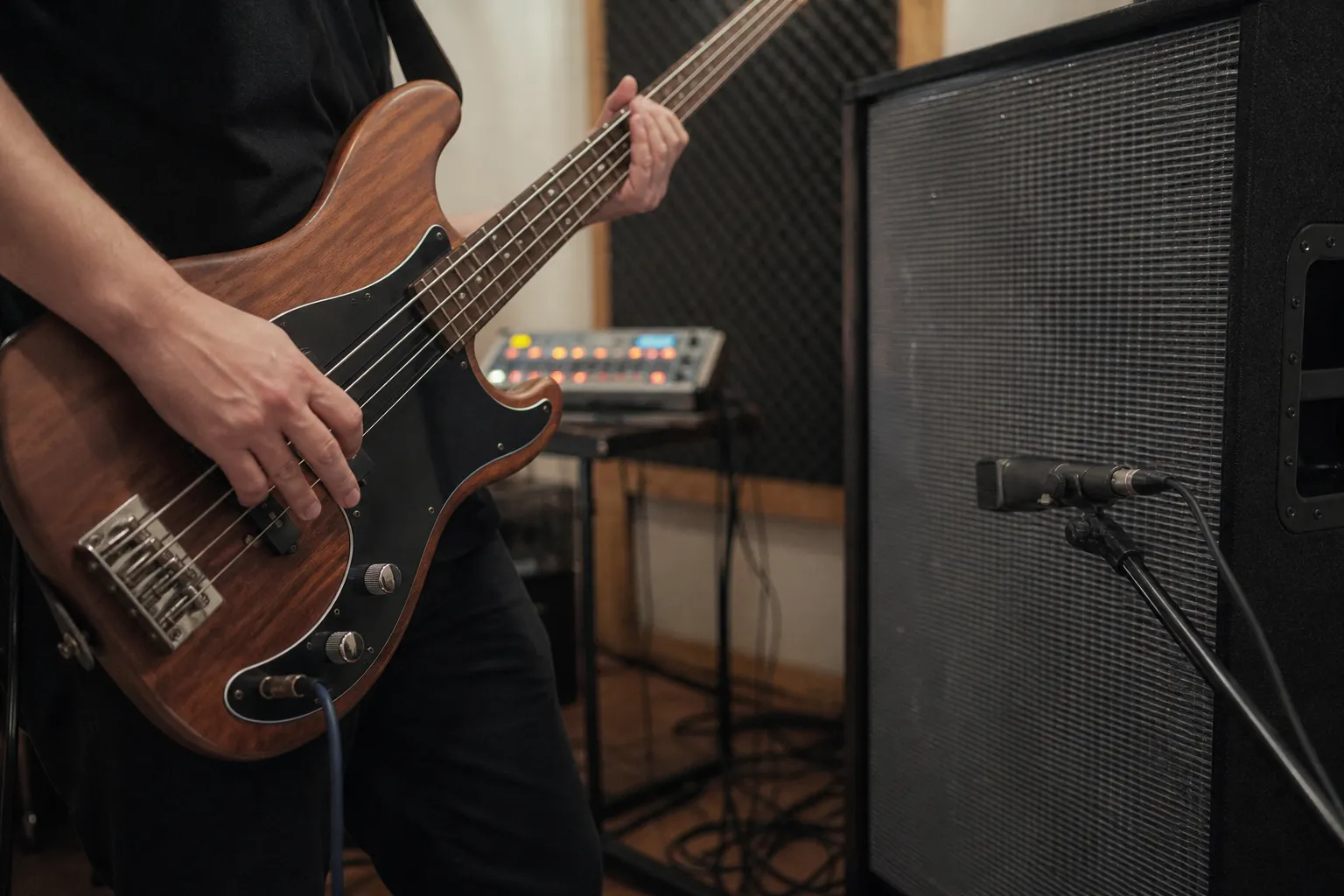 Conceptual editorial photograph showing an anonymous musician's hands playing bass beside a miked speaker cabinet, with a generic drum machine behind.