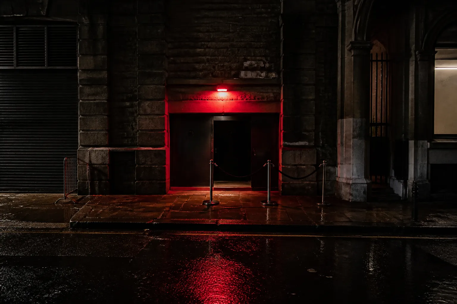 Conceptual editorial photograph of an anonymous narrow club entrance under a red light, with barriers and reflections on rain-wet pavement.