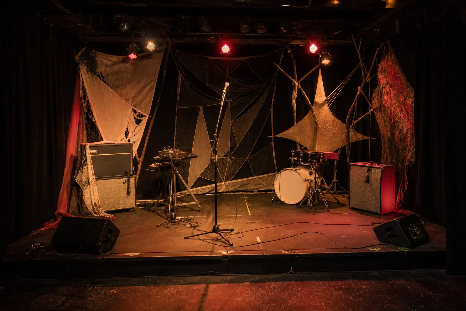 Conceptual editorial photograph of a tiny empty club stage with microphone, amplifiers, a compact machine stand, cables, black curtains, and handmade fabric decoration.