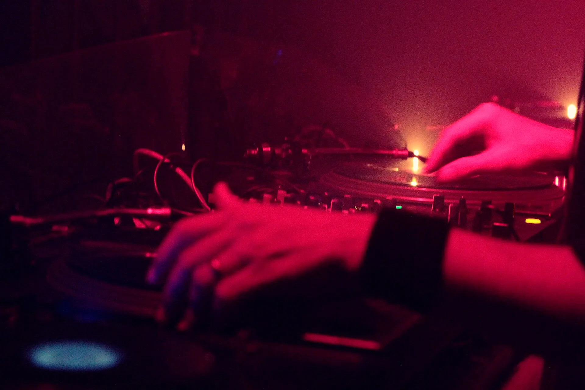 A club DJ wearing headphones reaches between two vinyl turntables and a central mixer while beatmatching records in a dark booth.