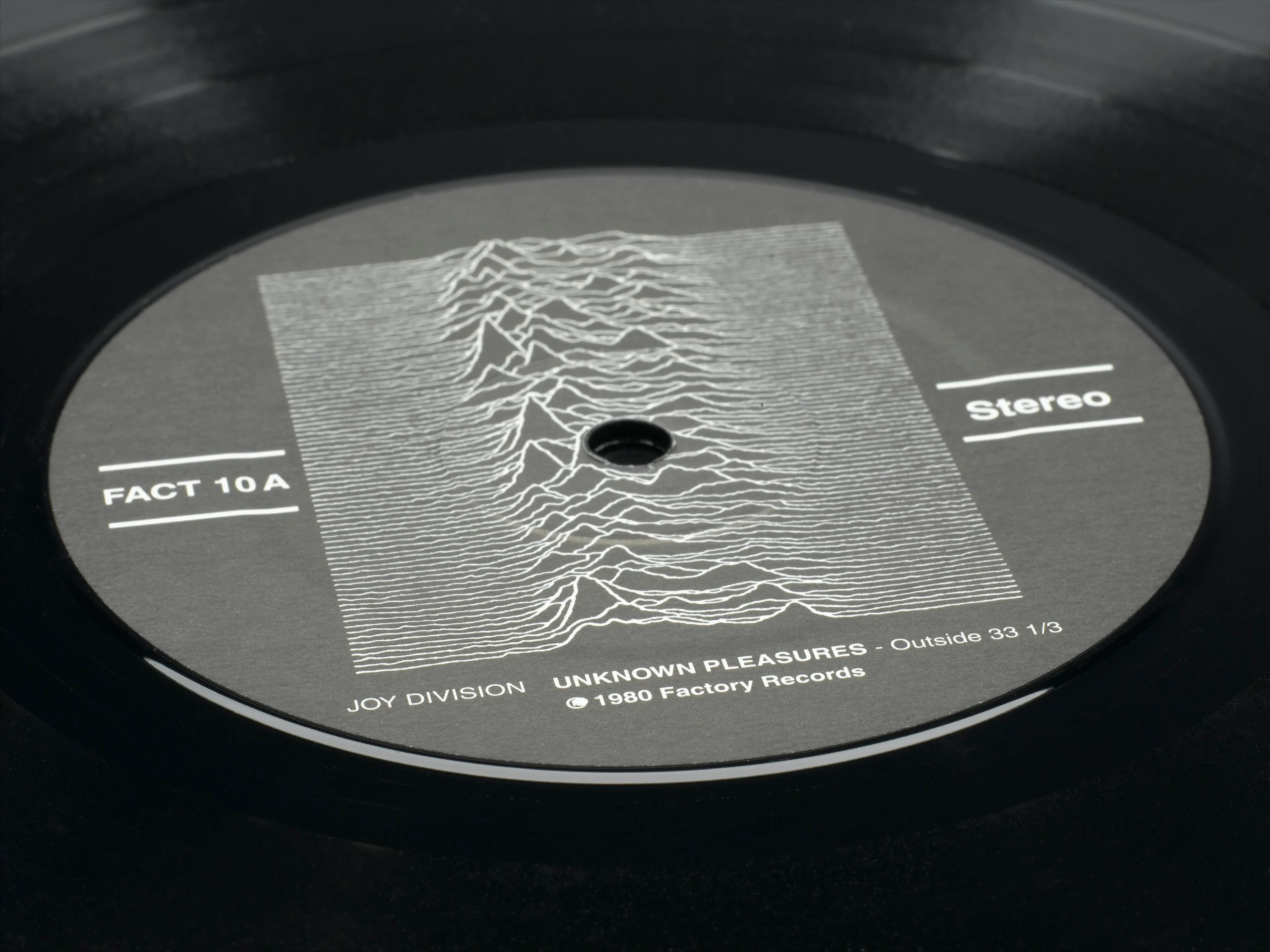 Close museum photograph of the Unknown Pleasures record label with the pulsar plot, FACT 10A number, and stereo marking.