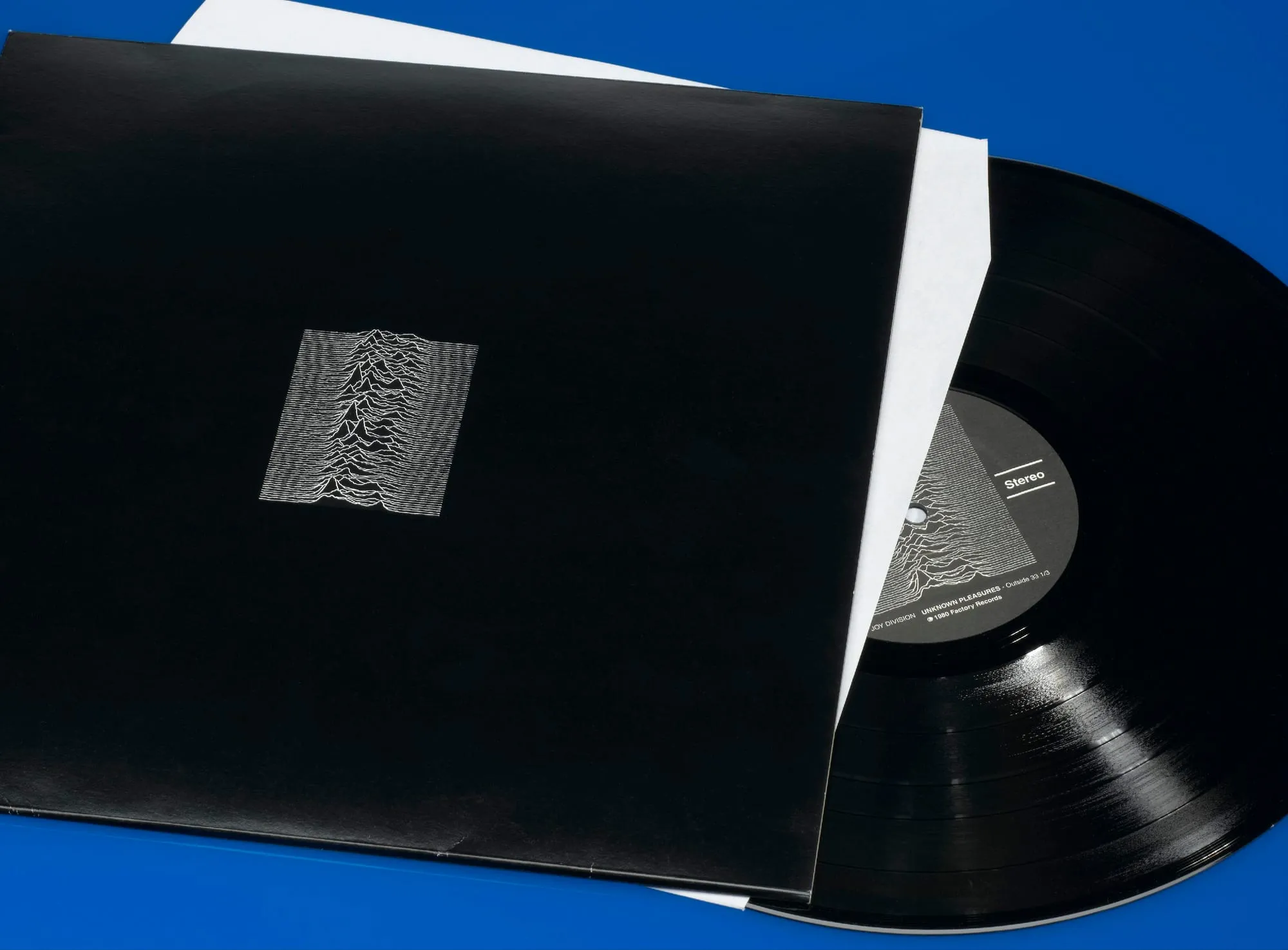 Angled museum photograph of the black Unknown Pleasures sleeve and partially removed record against a bright blue background.
